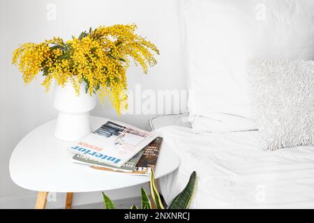 Vase mit Mimosenblumen und Zeitschriften auf Nachttisch in der Nähe einer hellen Wand Stockfoto