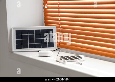 Tragbares Solarpaneel-Ladegerät, Mobiltelefon und Ohrhörer auf Fensterbank im Zimmer Stockfoto