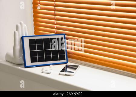 Tragbares Solarpaneel-Ladegerät, Mobiltelefon und Ohrhörer auf Fensterbank im Zimmer Stockfoto