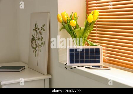 Tragbares Solarpaneel-Mobiltelefon und Vase mit Tulpen auf Fensterbank im Zimmer Stockfoto