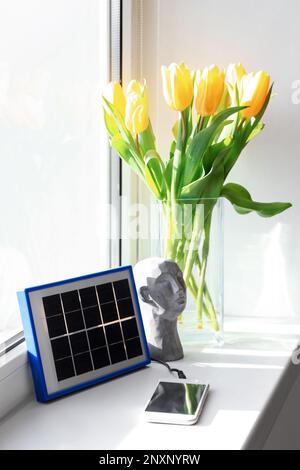Tragbares, mit Solarpaneelen ladbares Mobiltelefon, Dekor und Vase mit Tulpen auf Fensterbank im Zimmer Stockfoto