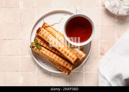 Platte mit Waferrollen mit abgekochter Kondensmilch und Tasse schwarzen Tee auf Fliesenhintergrund Stockfoto