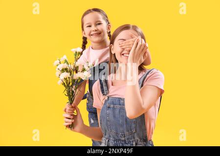 Ein kleines Mädchen schließt die Augen ihrer Mutter mit Blumen auf gelbem Hintergrund Stockfoto