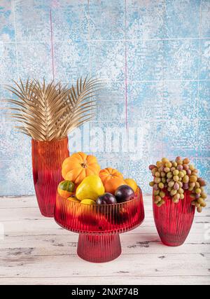 ZierKürbis, Obst und Trauben in einer Vase. Obsthalter für farbige Glasvase. Roter Glaskaraffe mit entferntem Deckel. Stockfoto