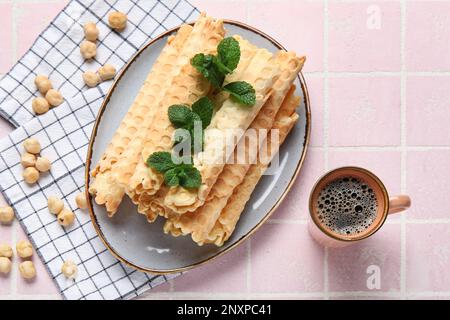 Teller mit köstlichen Waffeln, Minze, Haselnüssen und einer Tasse Kaffee auf pinkfarbenem Fliesenhintergrund Stockfoto