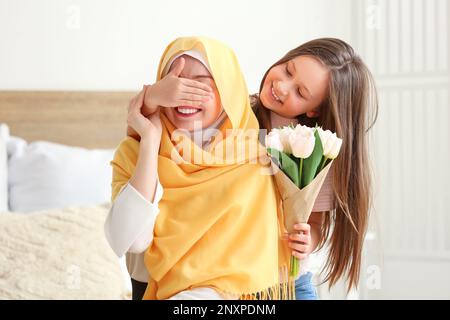 Ein kleines Mädchen mit Tulpen, das die Augen ihrer muslimischen Mutter im Schlafzimmer schließt Stockfoto