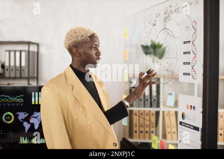 Junger afrikanischer männlicher Sprecher bei formellen Anlässen, der während einer Konferenz im Bürozimmer auf ein durchsichtiges Brett schreibt. Mitarbeiter des Unternehmens, der den Finanzbericht während der Geschäftsbesprechung präsentiert. Stockfoto