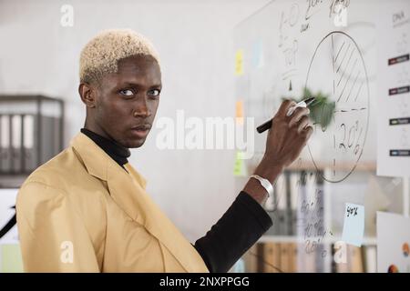 Selbstbewusster junger afrikanischer Mann in formeller Kleidung, der während eines Geschäftstreffens im Bürozimmer mit Papieraufklebern an Bord schreibt. Arbeitskonferenz- und Personenkonzept. Stockfoto