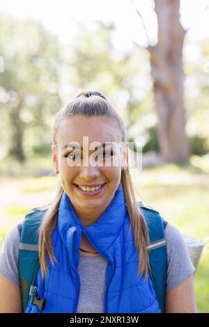 Vertikales Porträt einer attraktiven blonden Kaukasierin, die im sonnigen Wald wandert, lächelt, kopiert Stockfoto