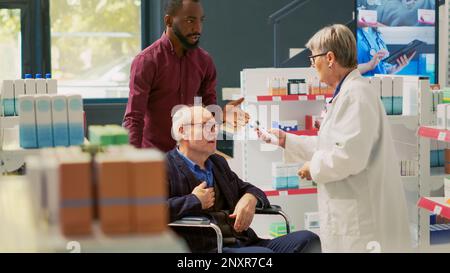 Spezialist zur Fiebermessung mit Thermometer bei älteren Menschen mit Behinderung, Rollstuhlfahrer und Sozialpflegepersonal. Kunde mit Behinderung im Gespräch mit Apotheker. Handgeführte Aufnahme. Stockfoto