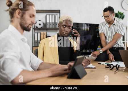 Fokussierter afrikaner spricht am Telefon, während seine männlichen Kollegen die Rede eines afroamerikanischen Geschäftsmanns hören. Firmenmanager, die im Büro arbeiten. Terminkonzept. Stockfoto
