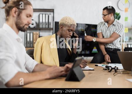 Fokussierter afrikaner spricht am Telefon, während seine männlichen Kollegen die Rede eines afroamerikanischen Geschäftsmanns hören. Firmenmanager, die im Büro arbeiten. Terminkonzept. Stockfoto