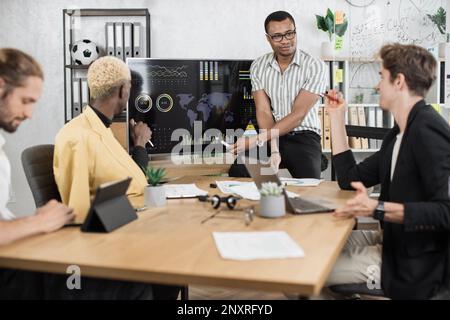 Fokussierter afrikaner spricht am Telefon, während seine männlichen Kollegen die Rede eines afroamerikanischen Geschäftsmanns hören. Firmenmanager, die im Büro arbeiten. Terminkonzept. Stockfoto