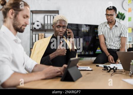 Fokussierter afrikaner spricht am Telefon, während seine männlichen Kollegen die Rede eines afroamerikanischen Geschäftsmanns hören. Firmenmanager, die im Büro arbeiten, zeigen Daumen nach oben. Terminkonzept. Stockfoto