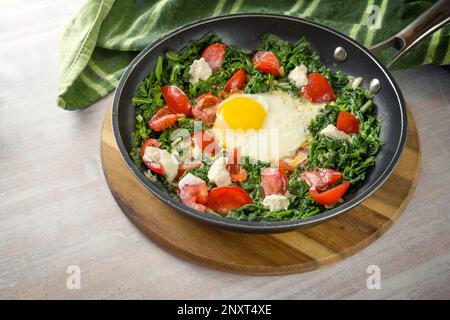 Spiegelei, Spinat und Tomaten mit Joghurt-Klumpen in einer Pfanne auf einem Holzbrett und einem grünen Küchentuch, gesundes Essen, Kopierraum, Stockfoto