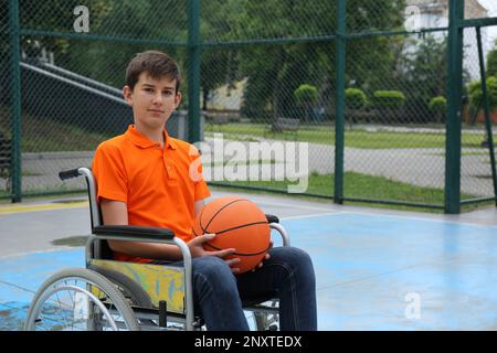 Behinderter Teenager im Rollstuhl mit Basketballball im Freien Stockfoto