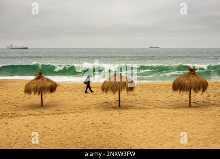 Ein einzelner Mann, der an einem kalten Sommertag an drei Palmenschirmen an einem leeren Strand in Valparaiso Chile vorbeigeht Stockfoto