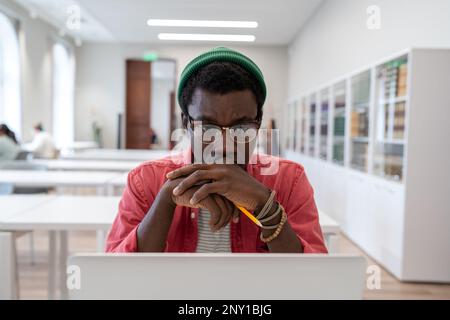 Rücksichtsvoller junger afroamerikanischer Freiberufler, der aus der Ferne an einem Laptop in einer öffentlichen Bibliothek arbeitet Stockfoto