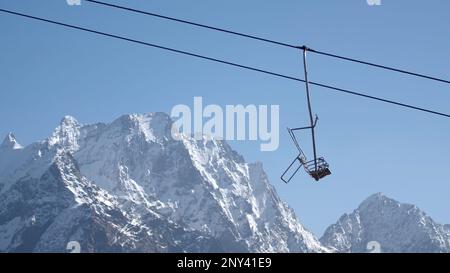 Seilbahn vor dem Hintergrund von Berggipfeln und blauem Himmel. Kreativ. Berglift mit beweglichen leeren Hütten mit schneebedeckten Bergen. Wunderschöne Landschaft Stockfoto