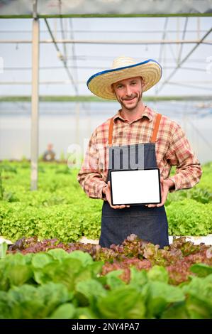 Ein glücklicher weißer männlicher Gärtner oder Farmer, der ein digitales Tablet mit einem weißen Bildschirm-Modell hält, während er im hydroponischen Gewächshaus steht. Stockfoto