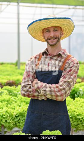 Porträt, erfolgreicher und glücklicher weißer männlicher Bauer mit Strohhut steht mit gekreuzten Armen in seinem Gewächshaus. Betriebskonzept Landwirtschaft Stockfoto