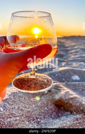 Die Sonne in einem Cognakglas am Strand. Hand mit einem Glas bei Sonnenuntergang. Stockfoto
