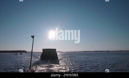 Blick auf die Fracht, die hinter dem Boot auf dem Fluss schwimmt. Clip. Der Frachtschiff fährt vor dem Hintergrund des sonnigen Himmels. Das Traktorboot zieht einen Lastkahn mit Fracht entlang des Ri Stockfoto