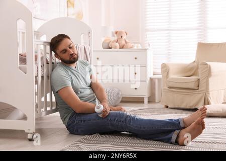 Müder Vater mit einer Flasche Milch schläft auf dem Boden im Kinderzimmer Stockfoto