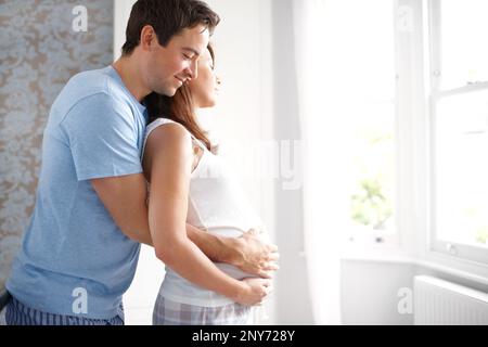 Unsere Träume werden wahr. Sie erwarten ein liebevolles Paar, das aus dem Schlafzimmerfenster schaut, während sie ihren schwangeren Bauch halten. Stockfoto