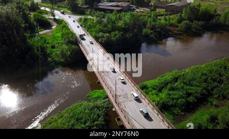 Luftaufnahme eines Flusses und einer Brücke mit Autos an einem Sommertag. Clip. Sommerdorf umgeben von grüner Vegetation Stockfoto