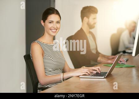 Jede Leistung beginnt mit der Entscheidung, es zu versuchen. Porträt einer jungen Designerin, die in einem Büro mit ihrem Kollegen sitzt, verschwommen im Hintergrund. Stockfoto