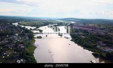 Luftaufnahme einer Sommerstadt, die von einem breiten Fluss durchquert wird. Clip. Brücke, die zwei Seiten einer Stadt verbindet Stockfoto