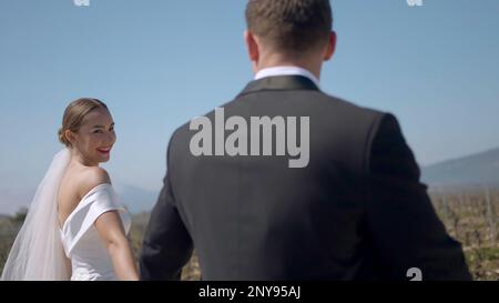 Glückliche Frischvermählte gehen Hand in Hand auf dem Feld. Aktion. Glückliche Frischvermählte, die an sonnigen Tagen auf dem Feld spazieren. Frisch Vermählte gehen gern auf dem Feld. Stockfoto