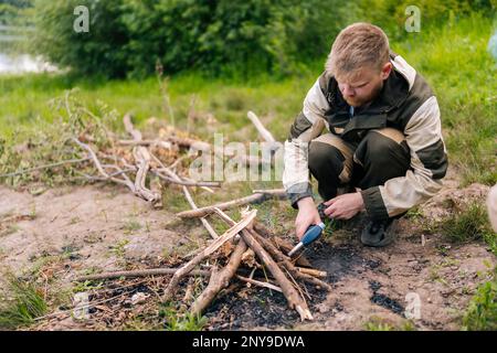 Bärtiger Überlebenskünstler in Regenjacke, der mit einem Gasanzünder am Flussufer Feuer zündet, um am Abend vor Sonnenuntergang zu kochen und sich zu erwärmen. Stockfoto