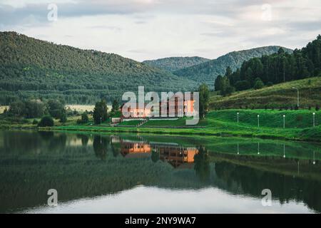 Wunderschöne Landschaft mit Hütte in der Nähe eines Sees und Bäumen. Holzhaus außerhalb der Stadt zur Erholung. Ökologisch saubere Region. Reflexion des Hauses Stockfoto