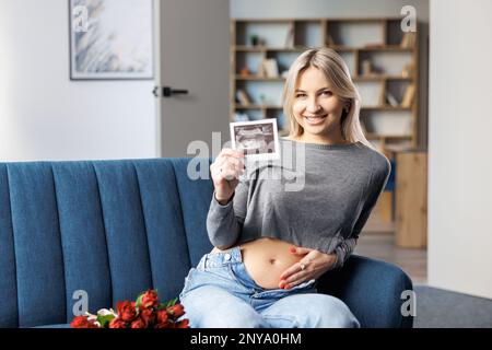 Foto einer schwangeren Frau zu Hause, die auf dem Sofa neben einem wunderschönen Blumenstrauß sitzt und in der Hand den Ultraschall-Scan-Druck des Babys hält Stockfoto