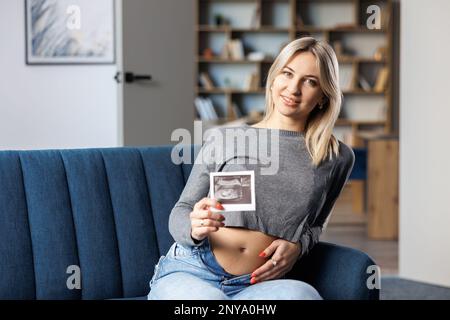 Foto einer schwangeren Frau zu Hause, die auf dem Sofa neben einem wunderschönen Blumenstrauß sitzt und in der Hand den Ultraschall-Scan-Druck des Babys hält Stockfoto