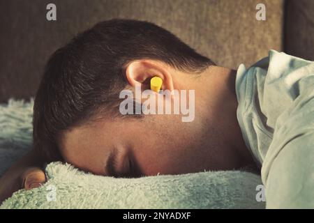 Mann mit Ohrstöpseln liegt im Bett. Tagsüber schlafen. Ruh dich aus, Siesta. Gelbe Ohrstöpsel in den Ohren eines kleinen Jungen, der nachmittags auf der Couch schläft, wenn p Stockfoto