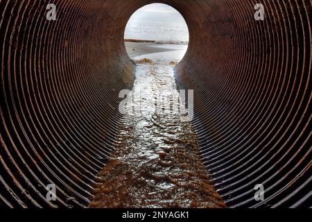 Blick von innen auf eine große rostige Abflussröhre, die zum Meer führt, mit einem Fluss aus Wasser und Sand flussabwärts zum Strand. Stockfoto