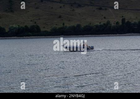 Ein Motorboot fährt mit hoher Geschwindigkeit auf dem Wasser. See mit felsigen Ufern, abendliche Landschaft. Stockfoto