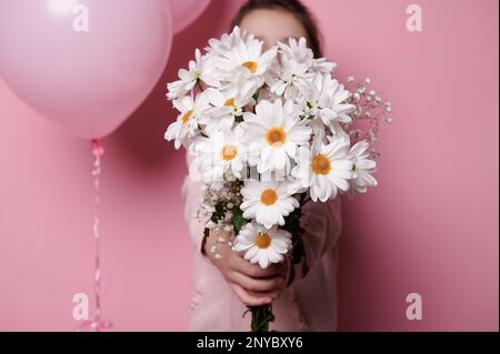 Ein Strauß weißer Kamillenblüten in Kinderhänden, isolierter rosa Hintergrund mit rosafarbenen Heliumballons in Pastelltönen Stockfoto