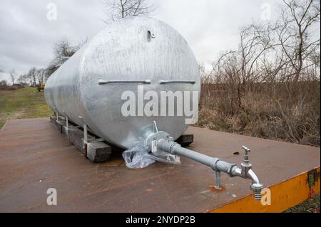 Silberner Metallwassertank mit Wasserpumpe vorne auf einem Anhänger im Freien neben einem landwirtschaftlichen Feld Stockfoto