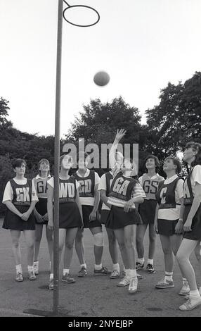 1989, netball, ein Schulmädchen-Team, mit Briefen auf ihren Westen, steht neben der Post, England, Großbritannien, einer der Mädchen (GA), die den Ball in den erhöhten Torring legt. Die Westen stehen für die sieben Positionen im Netball, d.h. GA-Goal-Angriff. Stockfoto