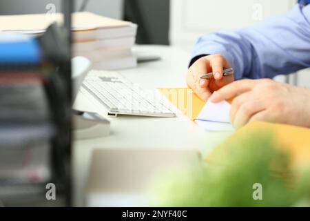 Ein Geschäftsmann sitzt am Tisch im Büro und hält einen gelben Umschlag in der Hand. Er nimmt ein Mitteilungsblatt für einen empfangenen Abschlussbrief aus Papier Stockfoto