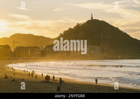 Leute spazieren am Sandstrand in der Nähe, winken dem Meer gegen Berge und Gebäude in der Abendzeit bei Sonnenuntergang Stockfoto