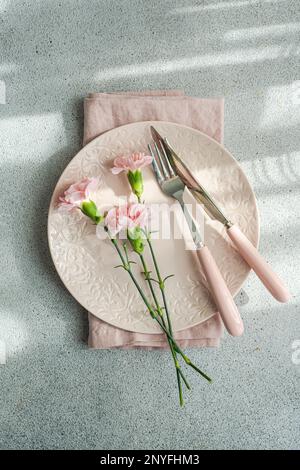 Von oben mit Teller und Serviette neben rosa Nelkenblumen auf Betontisch Stockfoto