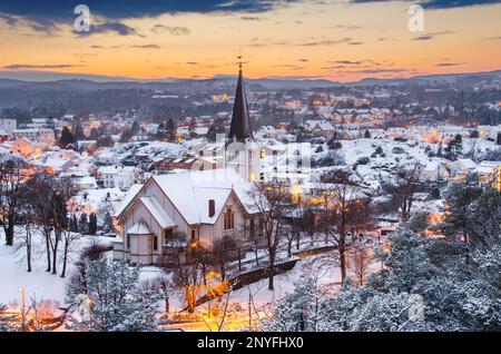 Malerischer Blick auf die lokale Kirche mit Wohnhäusern auf einem schneebedeckten Hügel vor dem bewölkten Himmel mit Winterwald in Grimstad Agder County Stockfoto