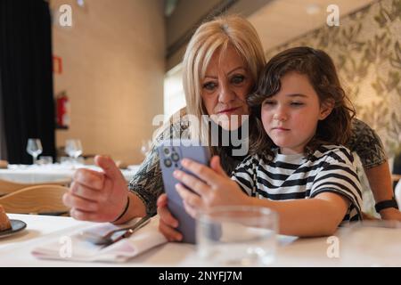 Großmutter und Enkelin sitzen am Tisch und machen Selfie auf dem Smartphone, während sie Zeit zusammen in einem modernen Restaurant verbringen Stockfoto
