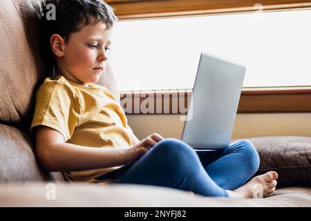 Konzentrierter kleiner Junge in lässigem Outfit, sitzt auf einem bequemen Ledersofa und surft im Wohnzimmer im Laptop Stockfoto