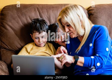 Positive, reife Großmutter lächelt, während sie auf einem bequemen Sofa mit niedlichen Kindern sitzt und zusammen mit dem Laptop surft Stockfoto